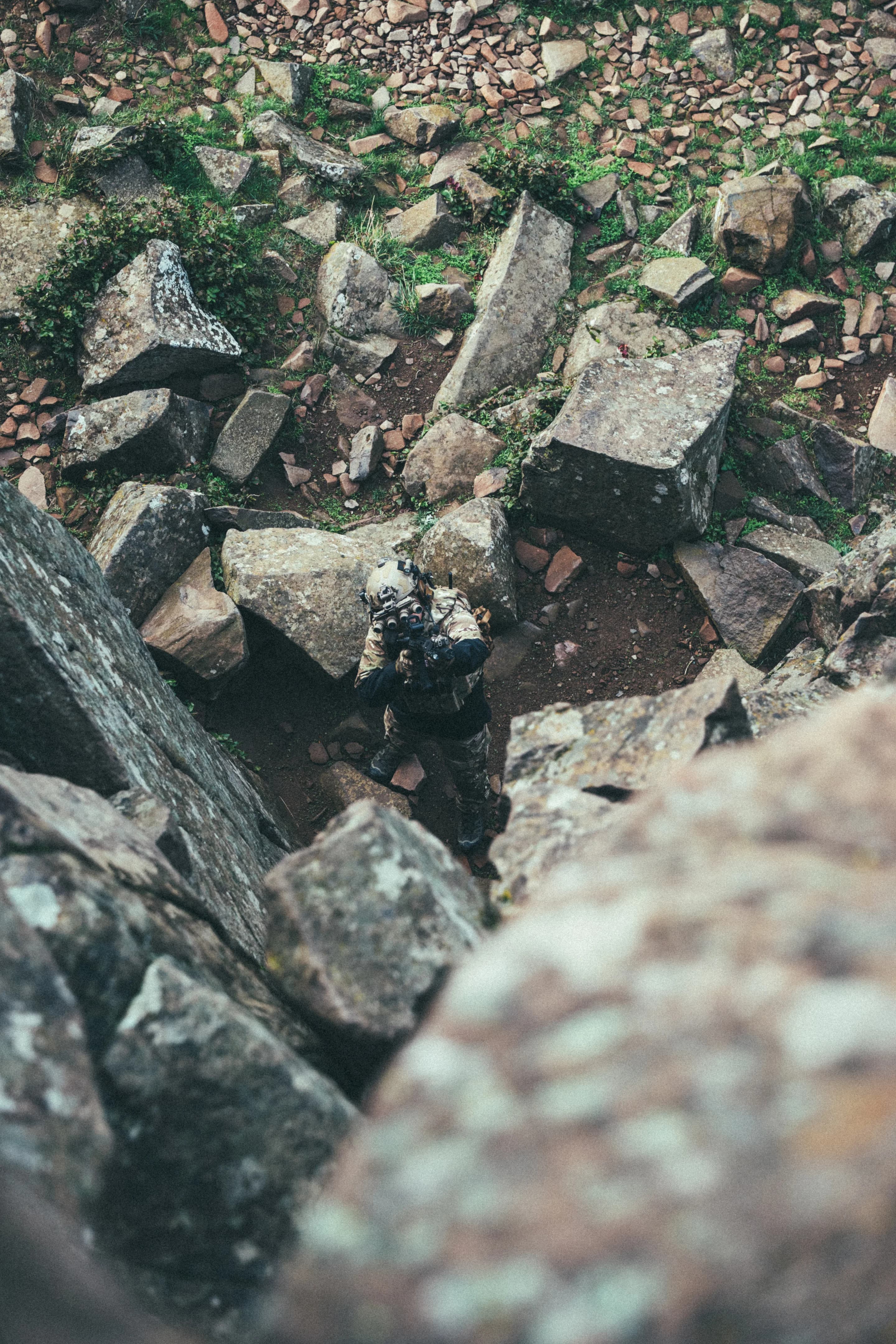 Operator aiming a suppressed rifle upward from a rocky crevice, seen from above